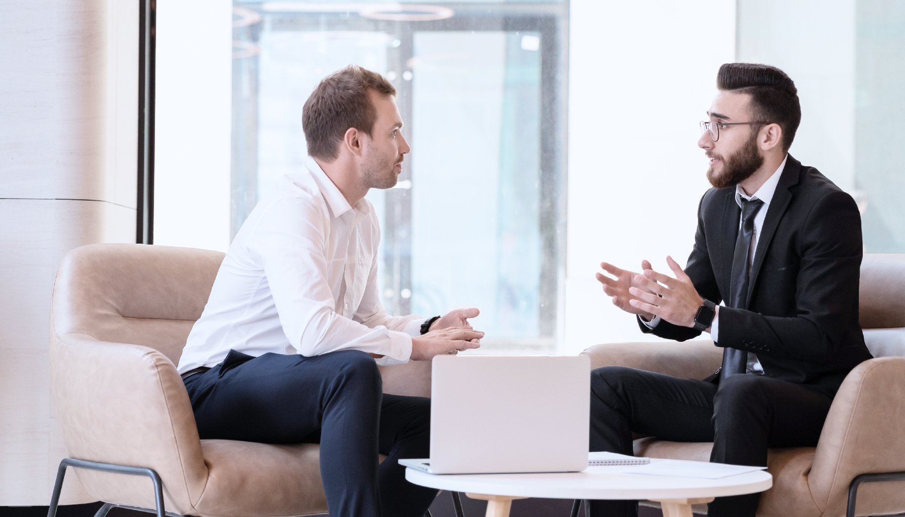 Two people sitting to have a conversation in an office with a laptop computer on a table between them.