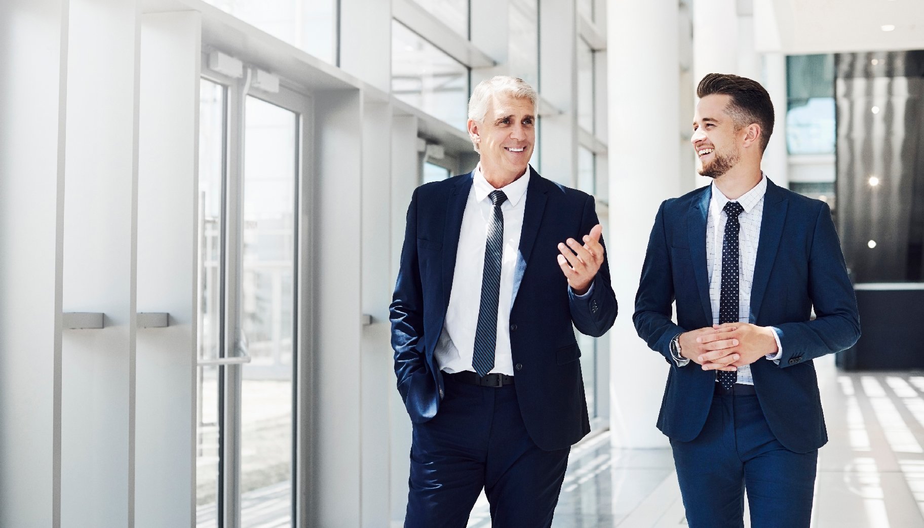 Two business people in dark blue suits talking in the hallway of a white office.